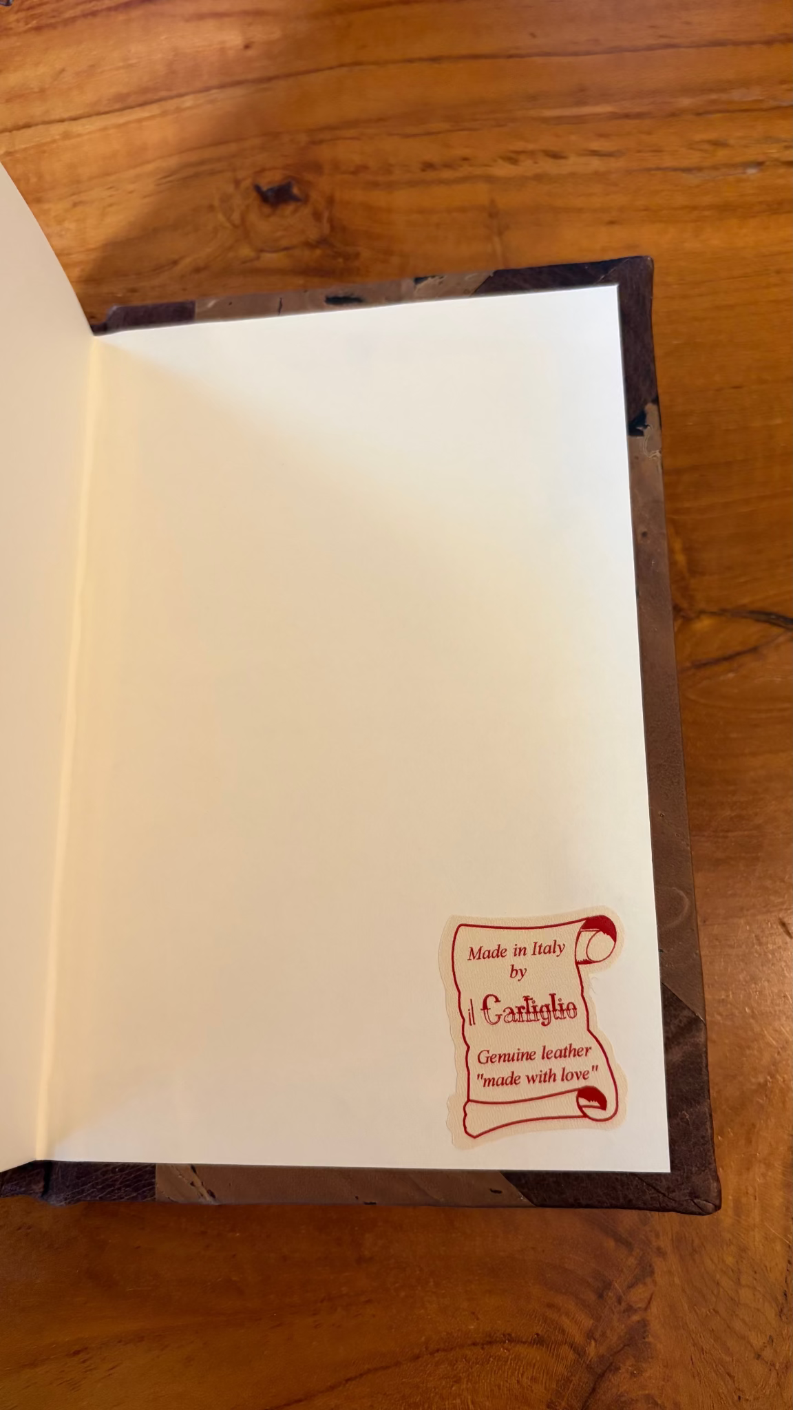 An open journal lying on a wooden table, showing blank cream-colored interior pages with a small red scroll-shaped label near the bottom right corner that reads 'Made in Italy by il Cartiglio'.