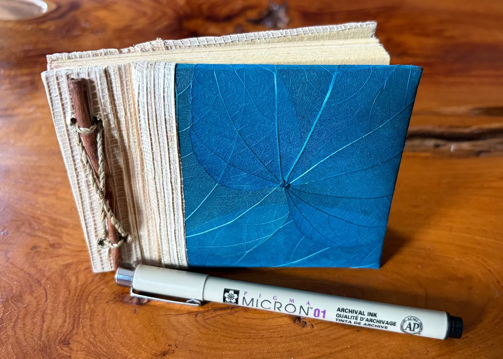 A handmade book or journal with a blue leaf-patterned cover and beige pages, standing on a wooden table next to a Micron pen.