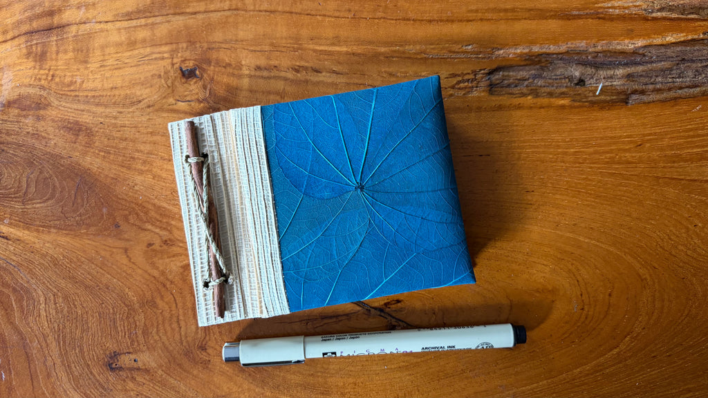 A square blue notebook with a leaf-like embossed cover and a striped fabric tie closure, placed on a wooden tabletop next to a pigma micron pen.