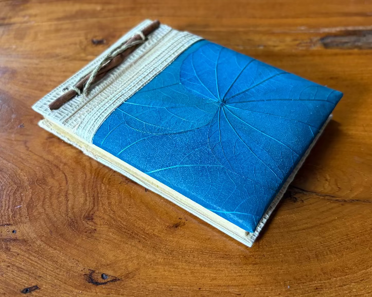 A blue handmade  journal with banana leaf embedded in the cover and a beige twine tie, resting on a rustic wooden table.
