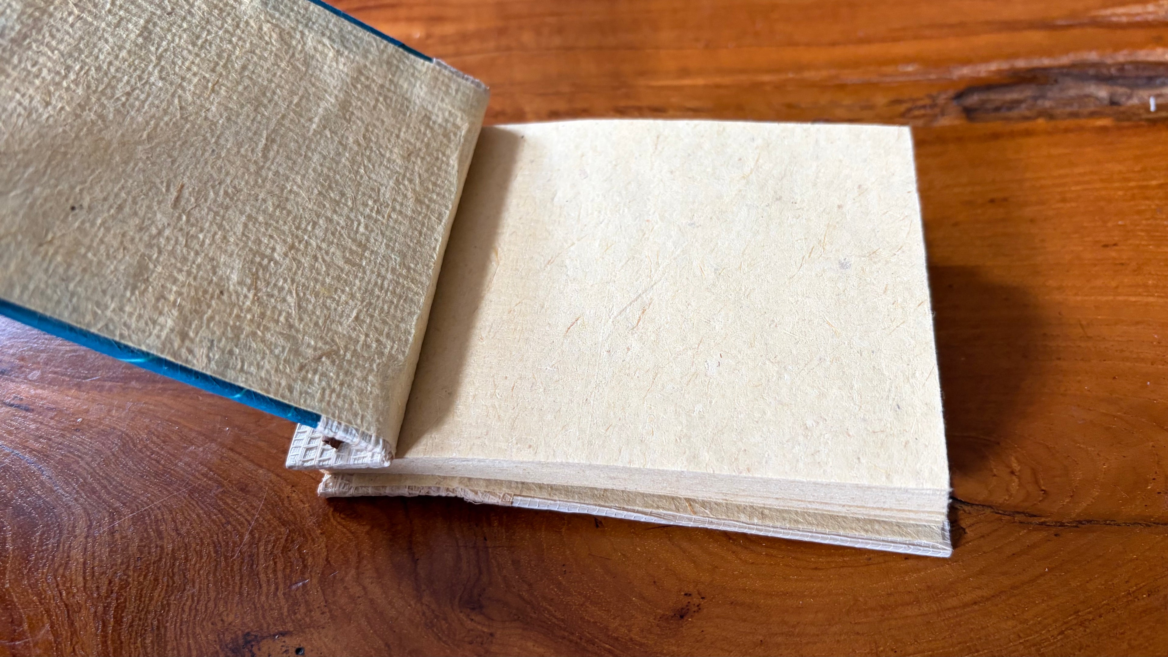 A handmade, square, blue notebook with a leaf-like embossed cover and a jute fabric spine, open on a wooden tabletop.