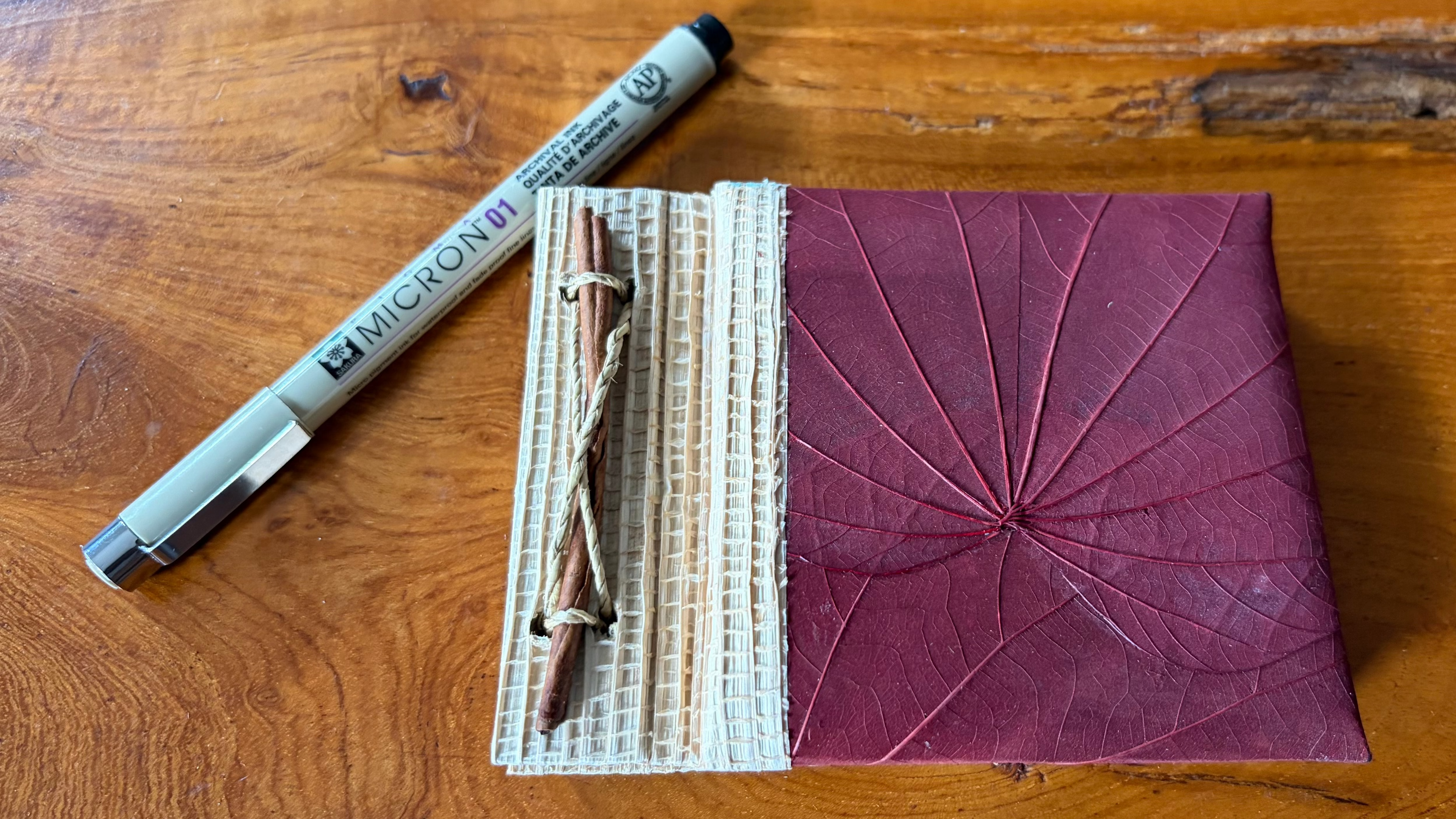 A handmade mini notebook with a textured cream paper cover and a deep burgundy,  leaf-vein cover, shown on a wooden table next to a Micron pen