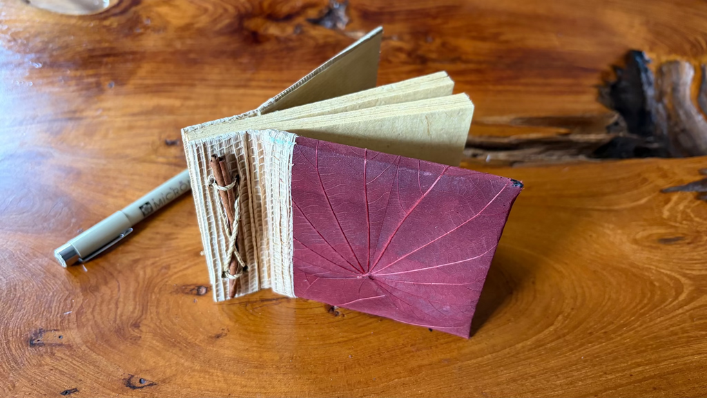 A handmade, square, burgundy mini art journal with a leaf-like embossed cover and a jute fabric spine, placed on a wooden tabletop next to a Pigma micron pen.