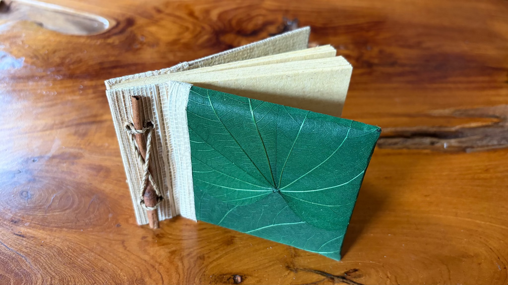 A small handmade journal with a green leaf-patterned cover and cream-colored pages, bound with a cinnamon stick and string, resting on a wooden table.