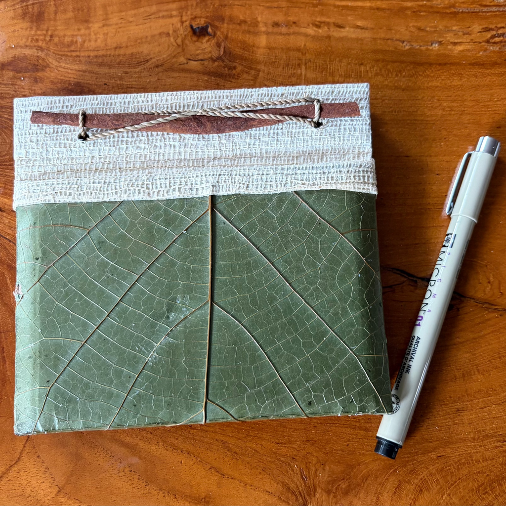 A handmade, square, green notebook with a leaf-like embossed cover and a jute fabric spine, placed on a wooden tabletop next to a Pigma micron pen.