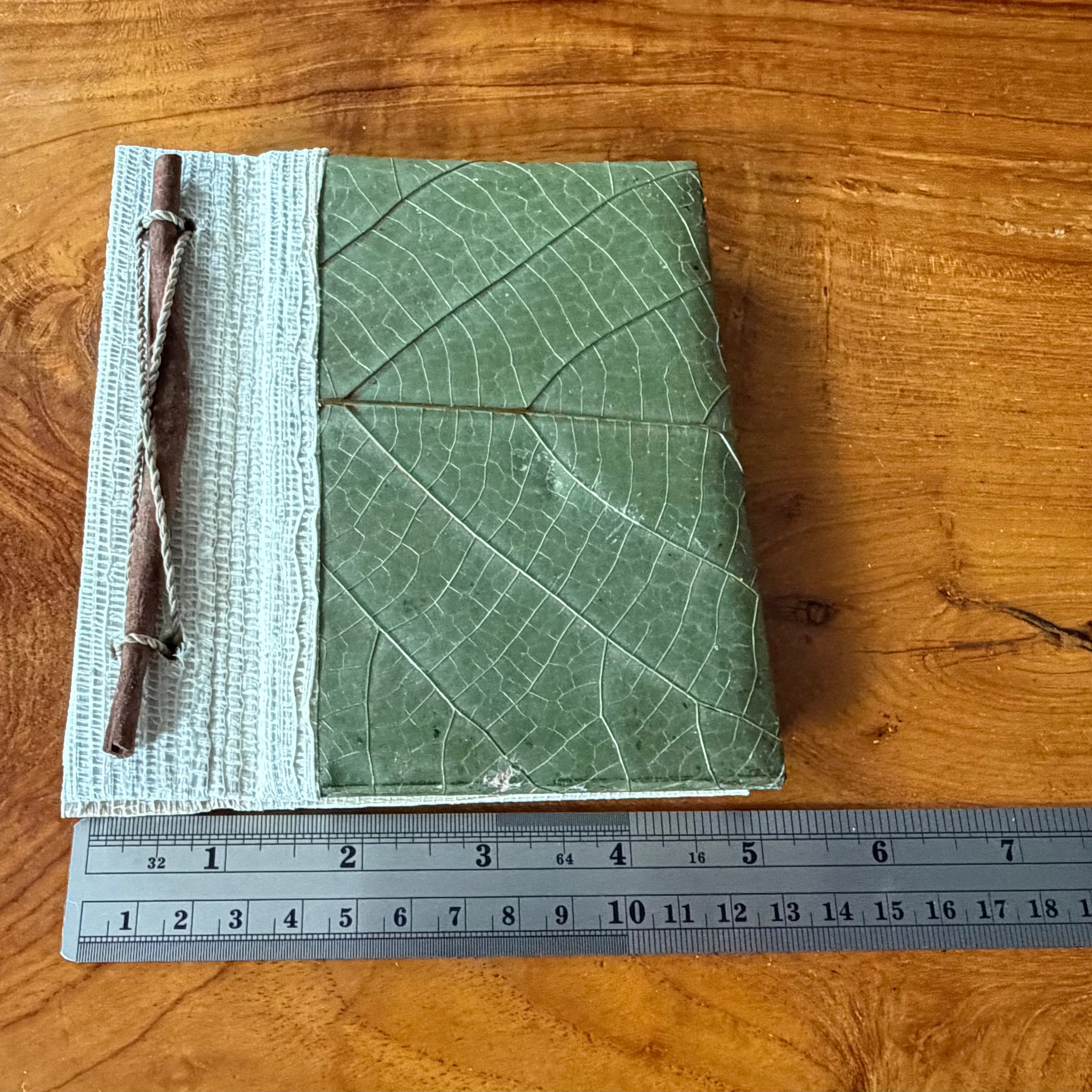 A square green notebook with a leaf-like embossed cover and a striped fabric tie closure, placed on a wooden tabletop next to a Pigma micron pen.