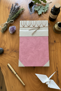 A pink journal or notebook with a textured cover, decorated with a cream band and twine, displayed on a wooden table surrounded by natural and craft props including dried lavender, amethyst, beads, a spoon, and a small bottle.